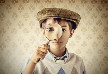 This is photo of a young boy wearing a Sherlock Holmes style hat. He is holding a magnifying glass to his eye, as though he is looking for clues.